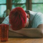 A man sits hunched over at a table with head down, covered by folded arms.