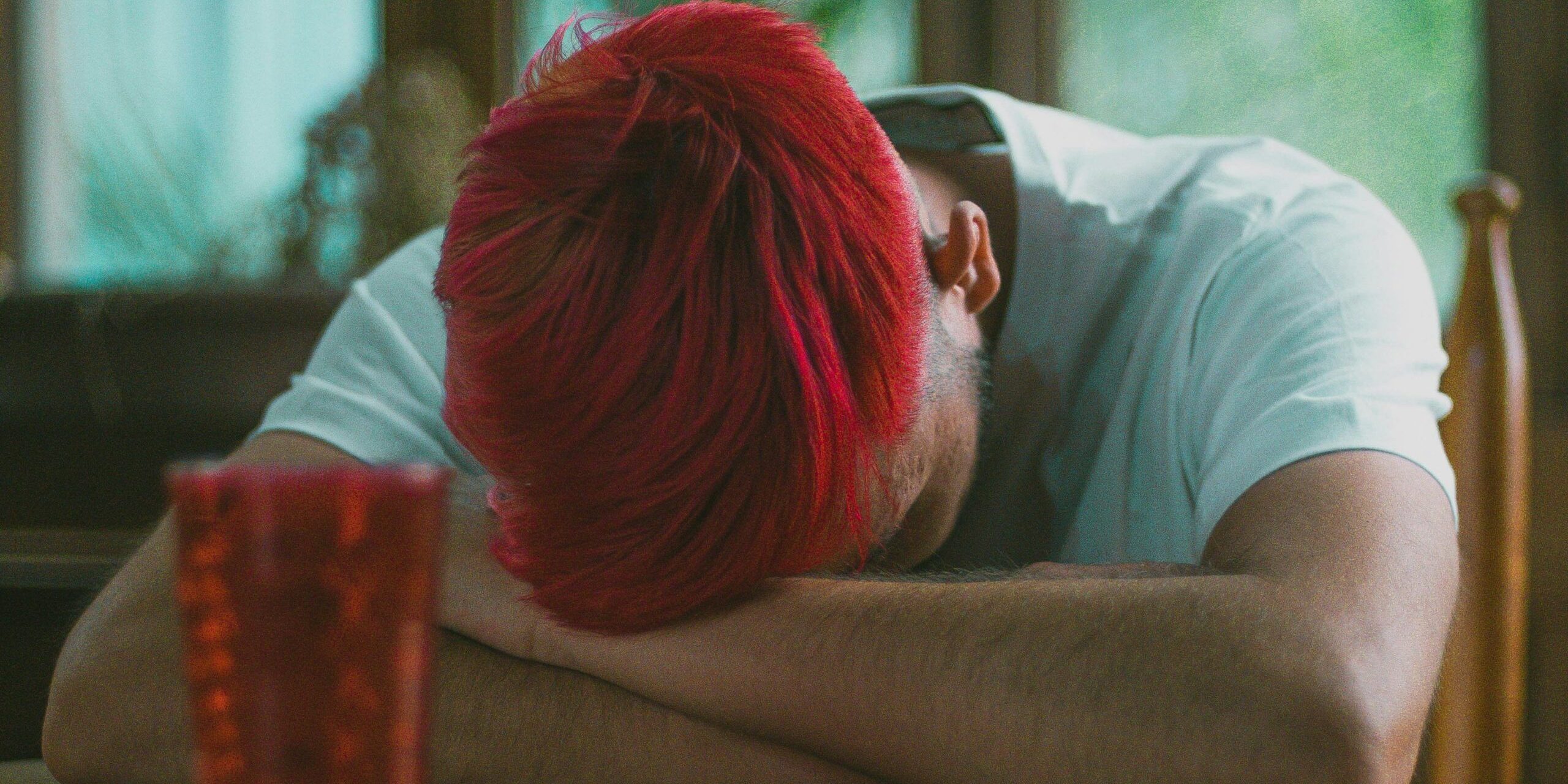 A man sits hunched over at a table with head down, covered by folded arms.